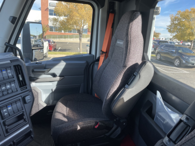 Interior view of a new Mack PR64T truck cab showing the driver's seat and dashboard. Seat has 'MACK' badge. Passenger window and door controls visible.