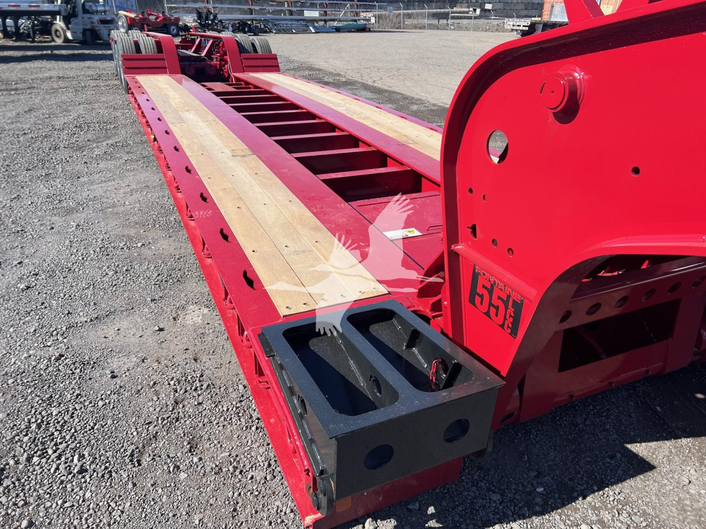 Lowboy trailer deck showing wooden planks and red frame with black tool/storage box at the front. 55E sticker and trailer framework visible.