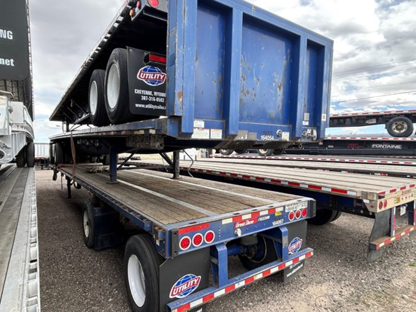 Flatbed trailer with a blue steel spread air bed on a trailer, tires visible, Utility sticker on back, spare tire on top rail, other trailers in background.
