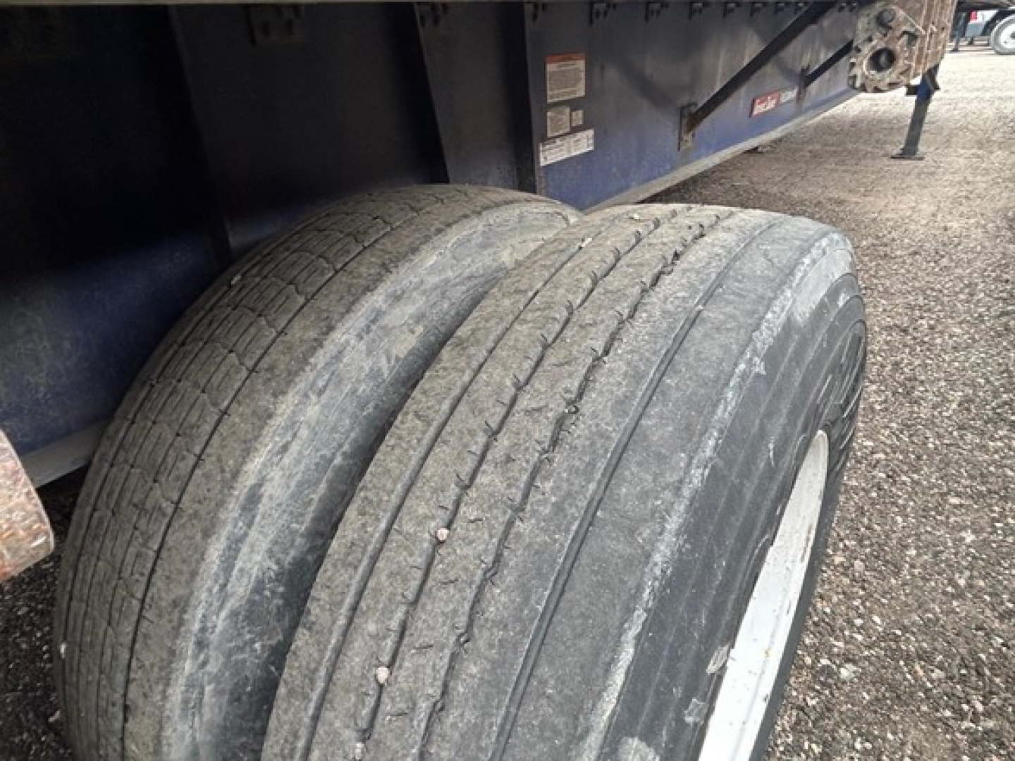 Dual rear tires on a flatbed trailer with wear bands; blue trailer frame and gravel ground visible.