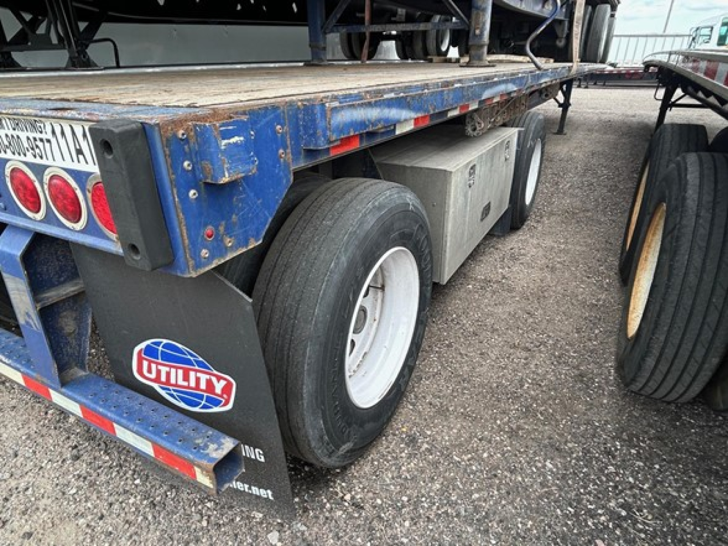 Flatbed trailer rear view with dual rear tires, blue frame, Utility logo mudflap, and rust-prone deck surface visible.