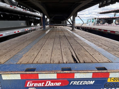 Flatbed trailer deck view from rear showing wooden planks and metal rim, Great Dane branding on rear edge, other trailers visible on sides.