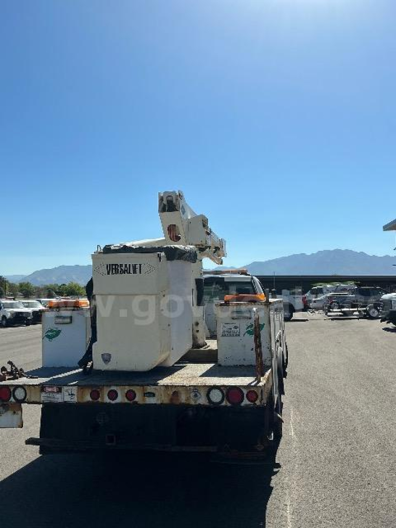 Rear view of a bucket truck on a flatbed with Versalift equipment, visible in a lot with mountains in the distance.