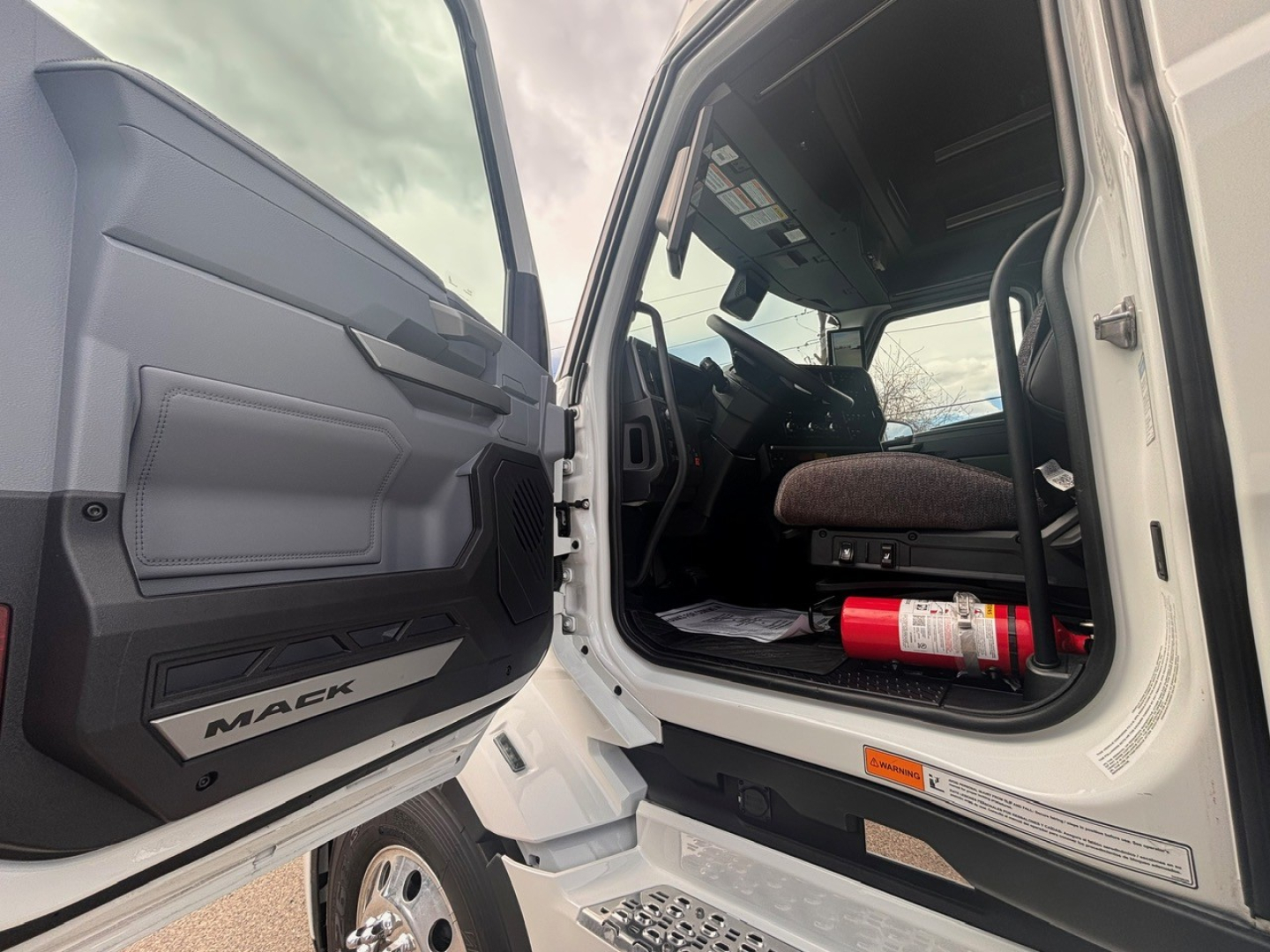 Mack PR64T76H conventional sleeper truck interior view from open driver door showing steering wheel, dashboard, seat, and a red fire extinguisher under the dash.