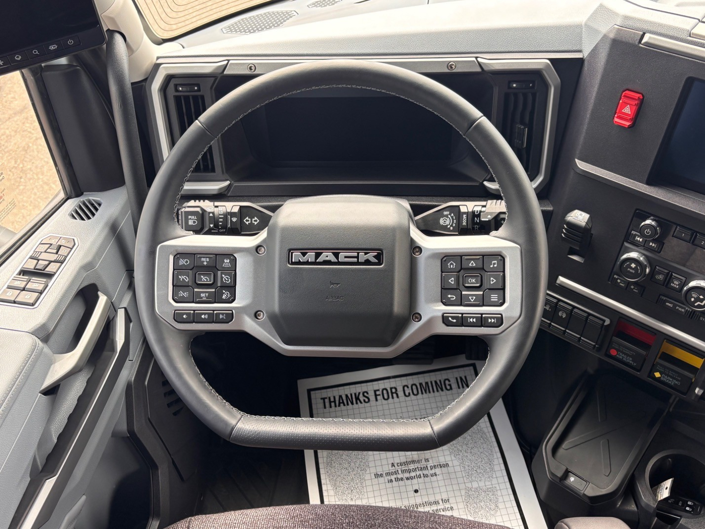 Steering wheel area of a Mack truck interior with multiple control buttons on either side of the central hub, dashboard vents, and a display screen. visible text includes MACK on the steering wheel center.