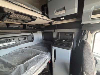 Truck cabin interior showing bunk bed with plastic-wrapped mattress, overhead storage, and compact fridge in a Mack PR64T76H sleeper.