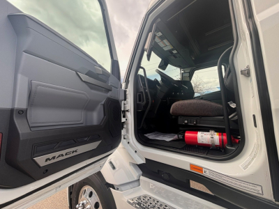 Mack PR64T76H conventional sleeper truck interior view from open driver door showing steering wheel, dashboard, seat, and a red fire extinguisher under the dash.