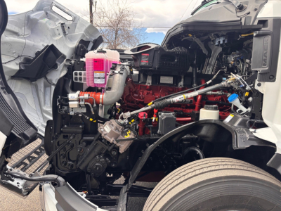 Truck engine bay with red engine, pink coolant reservoir, exposed hoses, and black chassis in a new Mack PR64T76H sleeper cab.