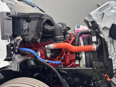 Engine bay of a 2026 Mack PR64T76H showing red engine, orange coolant hose, blue line, radiator, and surrounding components.