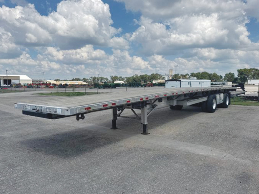 Flatbed trailer in a dealership lot showing long aluminum deck and tandem axles undercarriage with support legs.