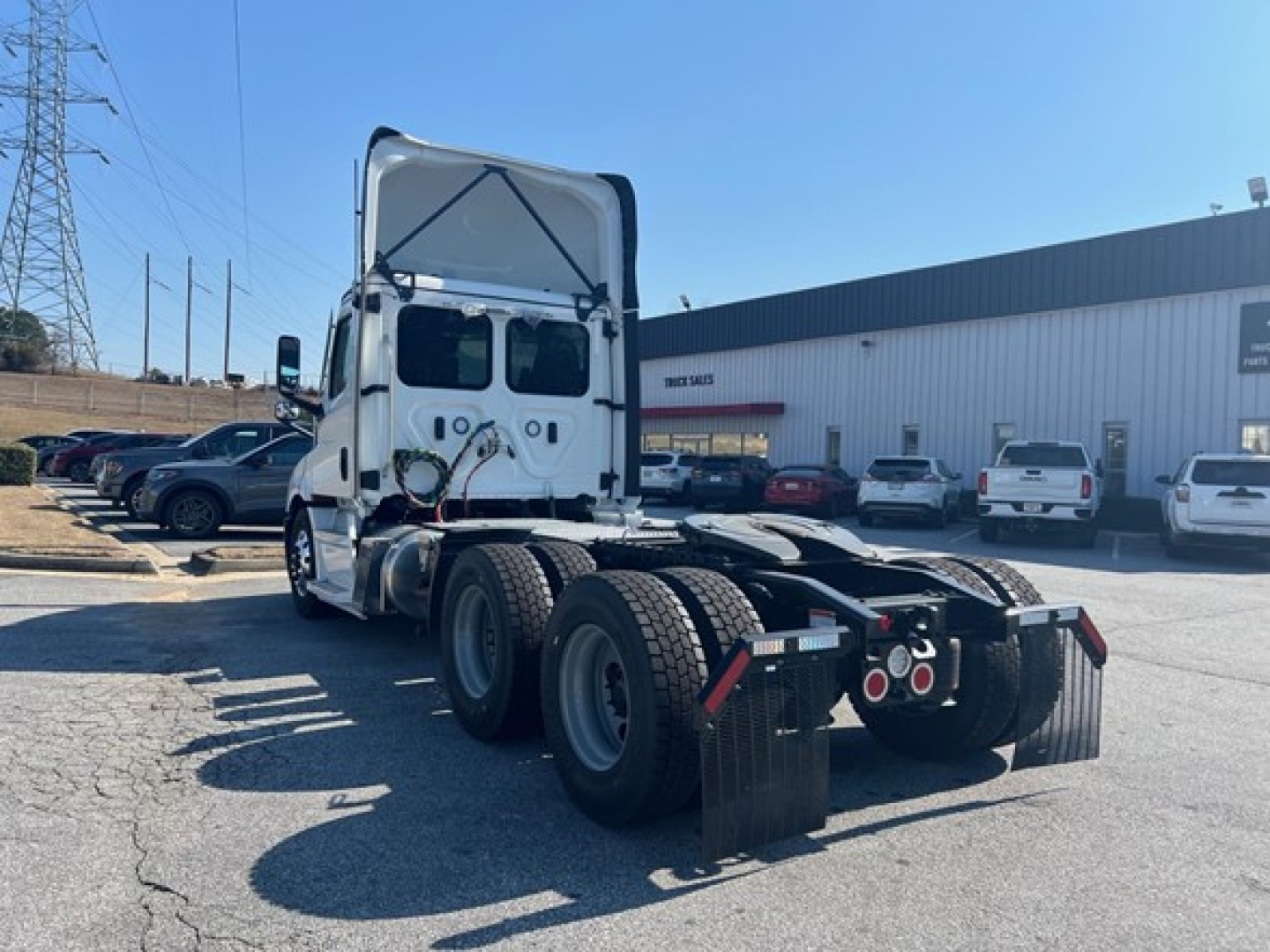 Freightliner Cascadia 116 day cab tractor with white cab, rear view showing sleeper area and fifth-wheel chassis, multiple parked vehicles in lot.