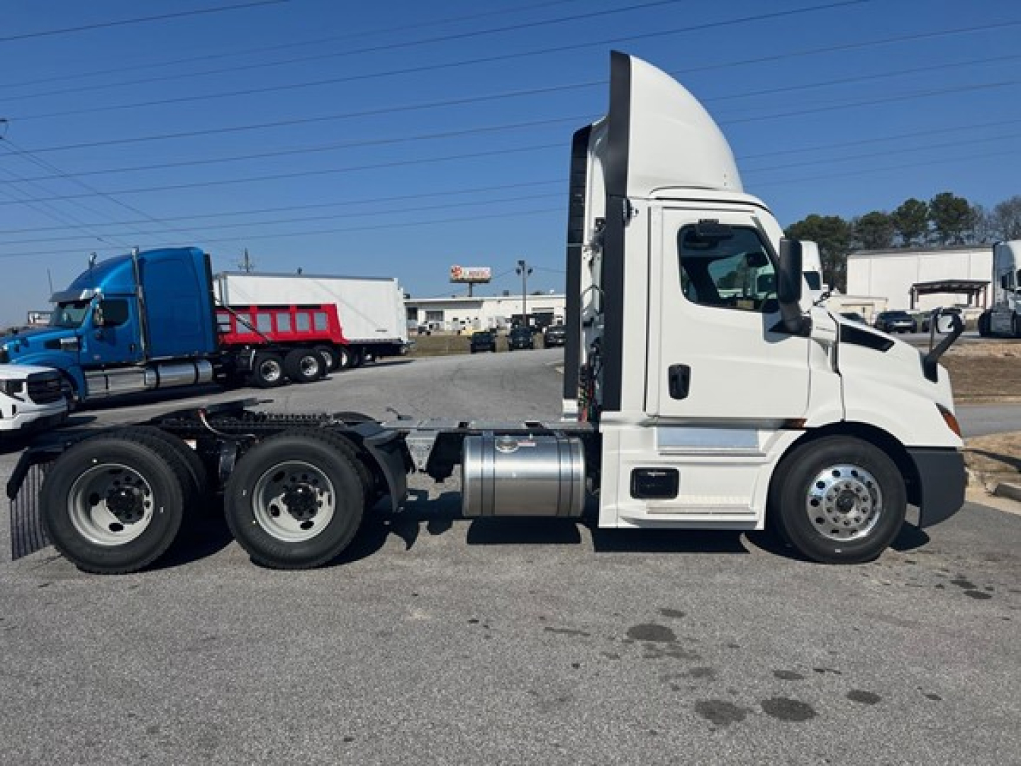 Two-axle tractor with white cab and blue trailer in background. Side view shows exhaust, fuel tanks, wheels.