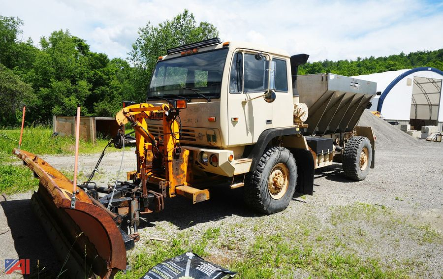 Military Plow & Spreader truck from 2003 Stewart & Stevenson with front plow attached, large dump bed and snow/ice equipment, white cab and tan chassis, on gravel lot.