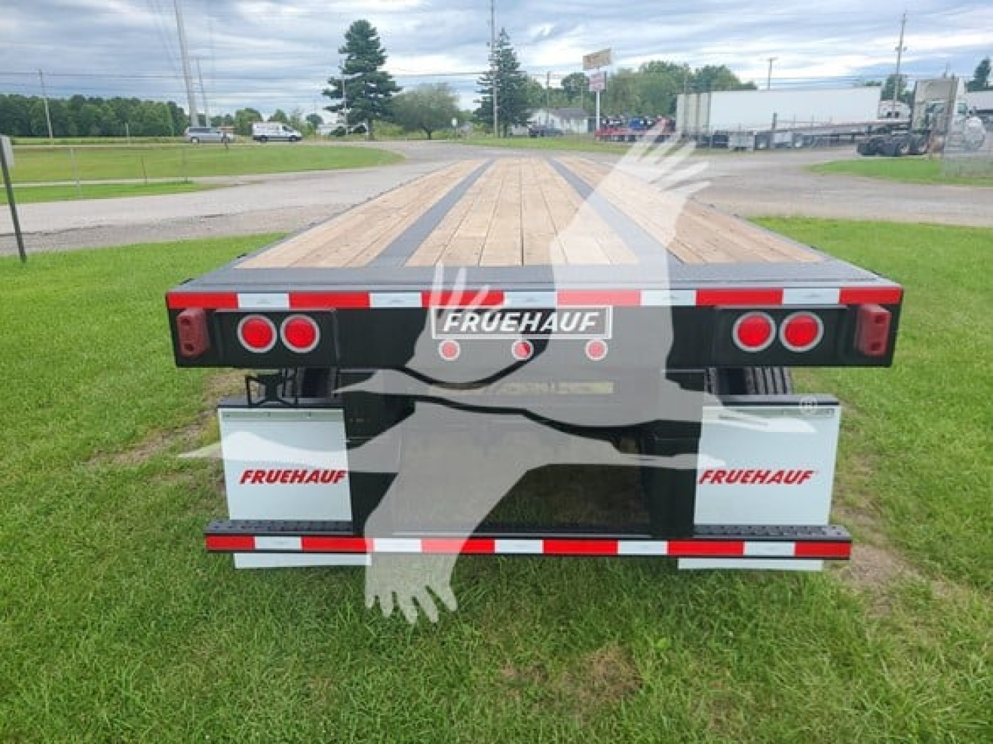 Flatbed trailer rear view showing FRUEHAUF branding, red rear lights, white FRUEHAUF boxes on each side, and wooden deck with black steel rails.