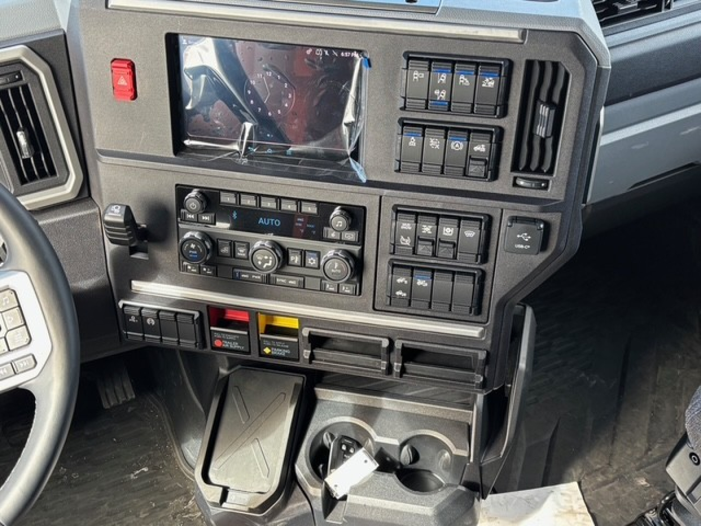 Dashboard of a 2026 Mack PR64T76H truck showing control panels, climate controls, and switches with a small display screen, cup holders, and gear area.