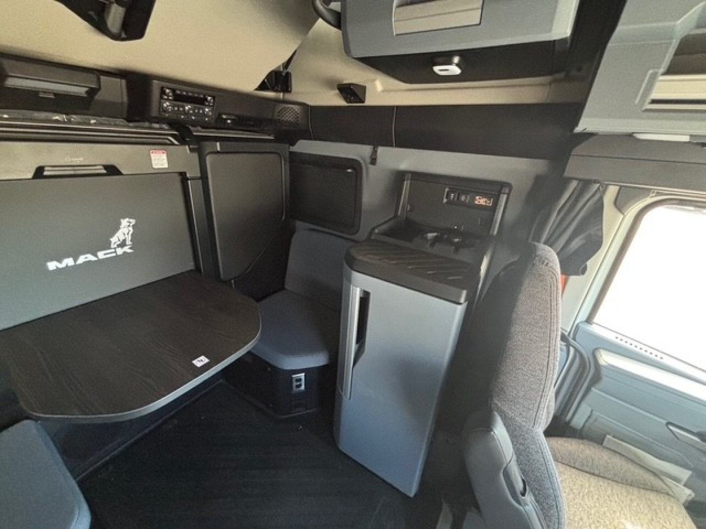 Interior of a Mack PR64T76H truck cab showing seating area, table with Mack logo, small fridge, storage compartments, and dashboard panels.