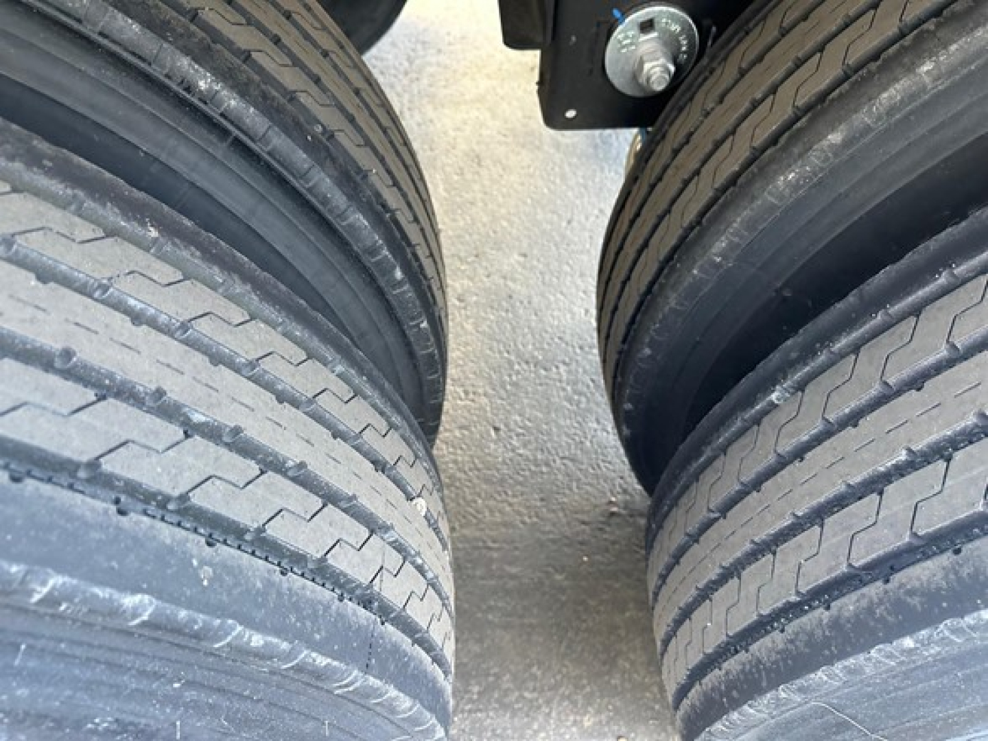Trailer axle view showing two dual tires and part of the suspension; close-up of tread pattern on tires and a scatter of gravel beneath.