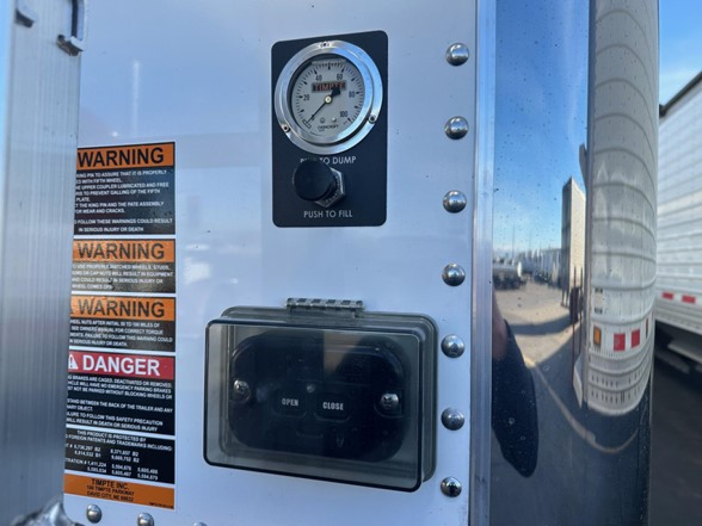 Panel on trailer showing a pressure gauge labeled 'DUMP' with scale, a push-to-fill label, a rectangular access door with 'OPEN' and 'CLOSE' indicators, and stacked warning labels on the left.