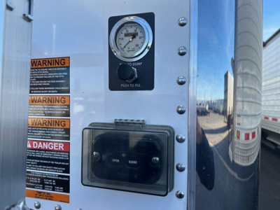 Trailer gauge panel showing a circular pressure gauge and a square door labeled OPEN/CLOSE, with warning stickers on the left and a reflection of trucks on the right.