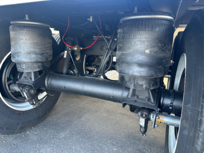 Hopper trailer undercarriage showing air suspension bags, axle, and drivetrain components. Visible text on bags includes ‘KENWORTH’ style branding; no legible serials or part numbers readable.