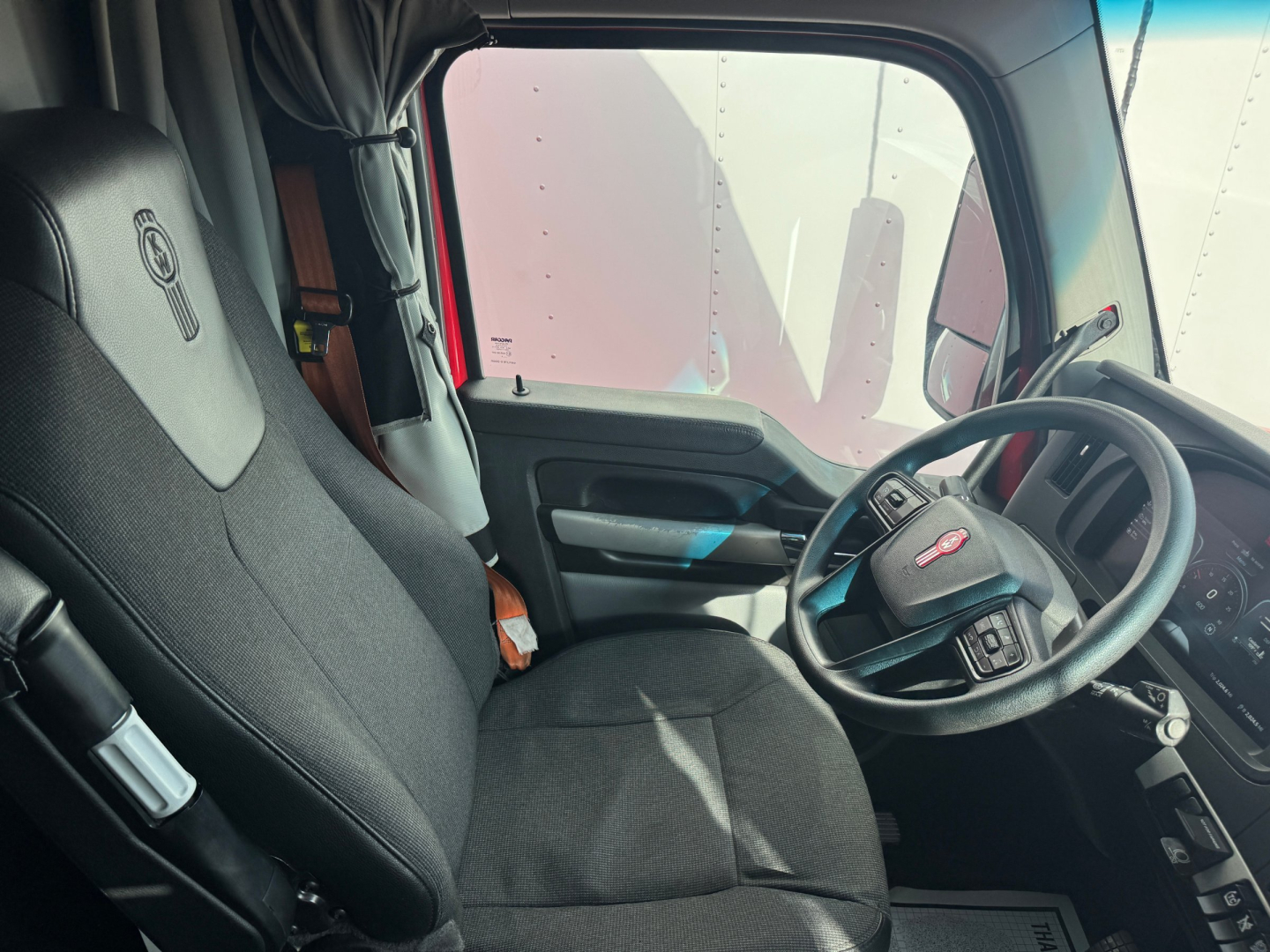 Kenworth T680 interior view showing driver seat, steering wheel, and dashboard; fabric seats with Kenworth logo on headrest.