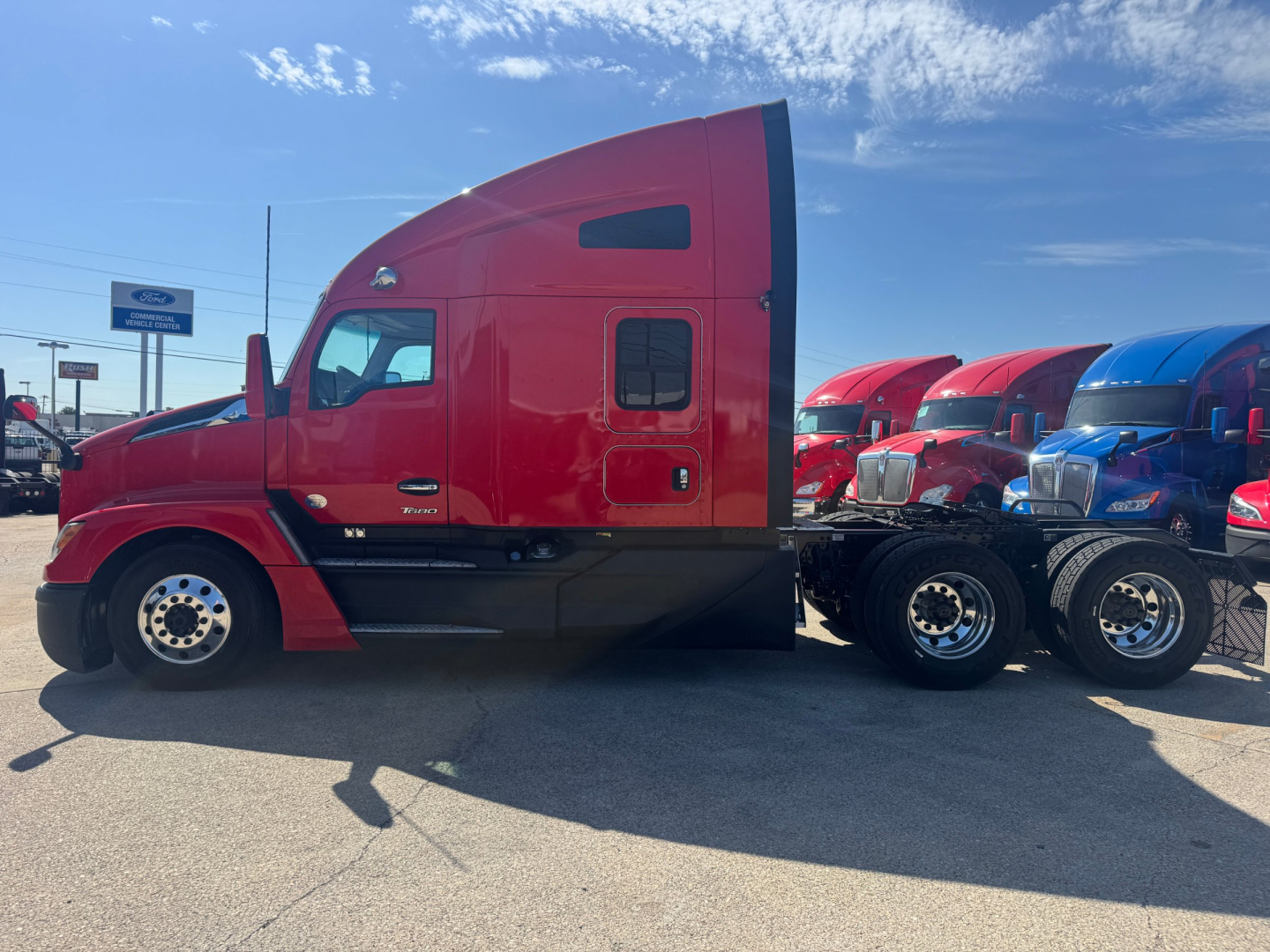 Kenworth T680 semi-truck side profile in red with sleeper cab, black chassis, and multiple trucks parked in background.