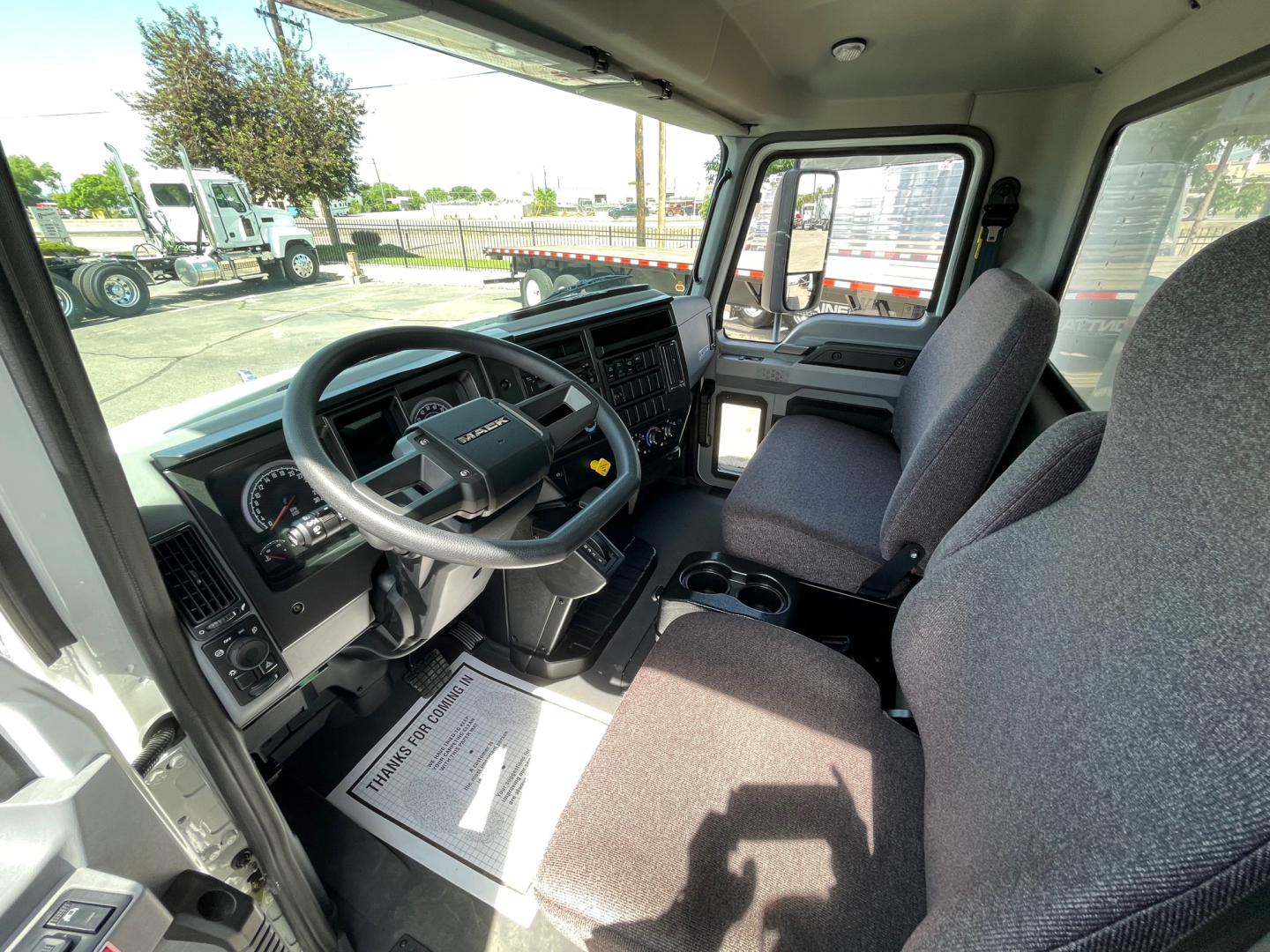 Mack MD6 truck interior cab view showing steering wheel, dashboard, and driver seat; front passenger seat visible; window and exterior trucks in background.