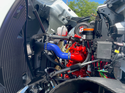 Mack MD6 engine bay with blue intake hose, red engine block, black electricals, and various hoses and reservoirs visible.