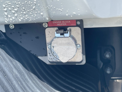 Engine block heater access cover with red label reading ENGINE BLOCK HEATER on a Mack MD6 truck engine bay.