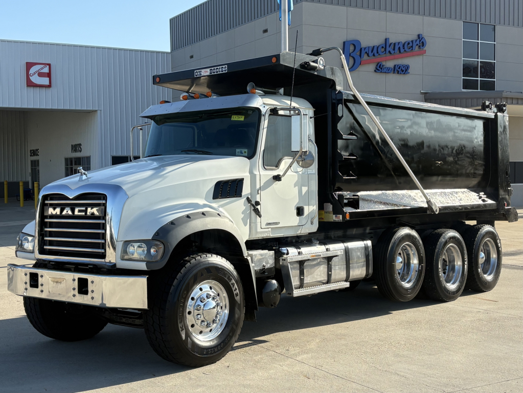 Dump truck: 2021 Mack GR64F in-service white cab, black dump body, front grille with MACK letters, chrome bumper, dual rear axles, and heavy-duty wheels; dealership lot in background.