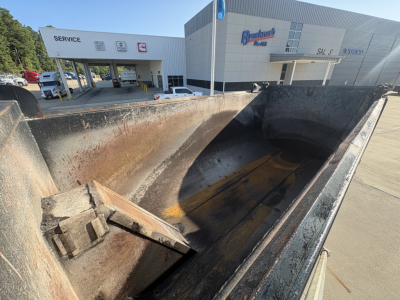 Dump truck bed interior of a 2021 Mack GR64F showing worn metal walls and a rust-stained bed with some wooden blocks; dealership buildings in background.