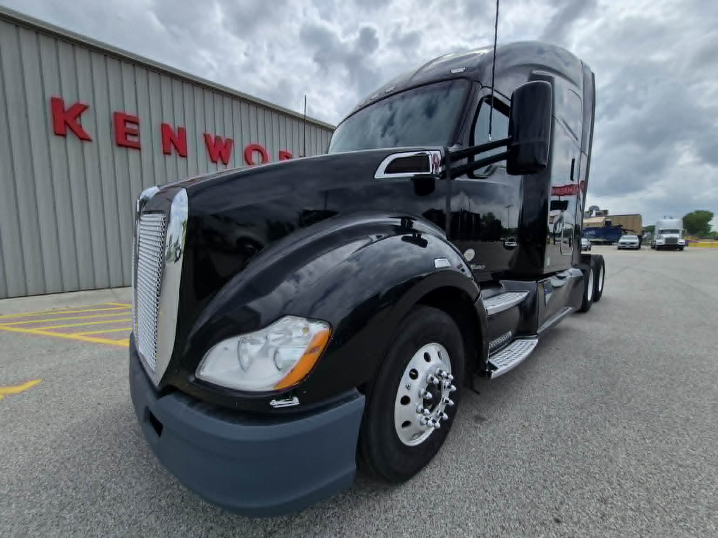 Kenworth T680 driver-side front view showing black tractor in a truck yard with Kenworth building in background.