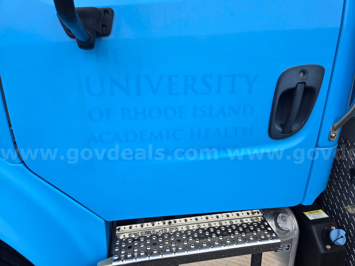 Truck door with blue paint showing partial text from a watermark: 'UNIVERSITY OF RHODE ISLAND ACADEMIC HEALTH' and website 'www.govdeals.com' along with a metal textured step and round headlight partially visible.