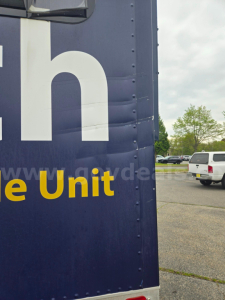 Truck trailer side with partial large white letters and yellow 'Unit' text, blue background, dealership lot visible in distance.