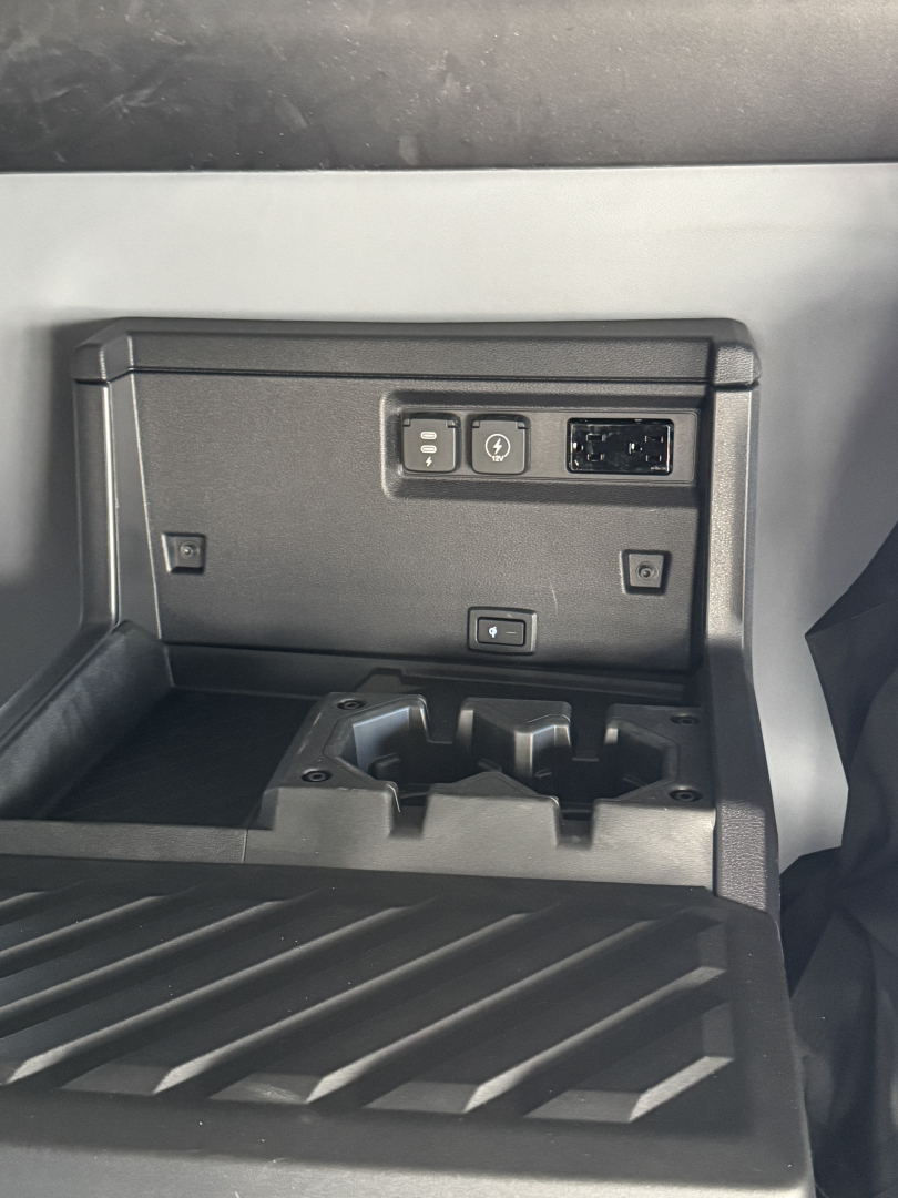 In-cab storage with three rectangular controls on a panel and a small USB/12V port, plus a stepped rubber floor mat.