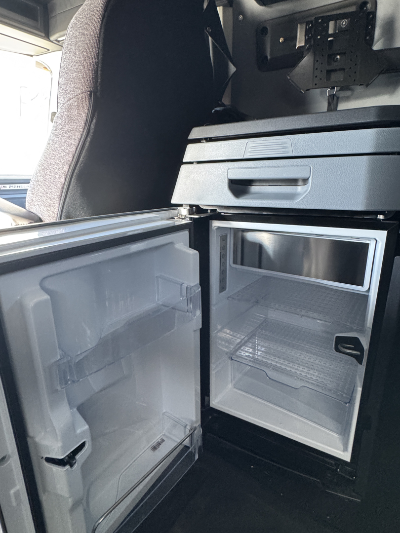 Truck cab interior showing a compact under-dash refrigerator/freezer unit with open double doors and empty shelves.