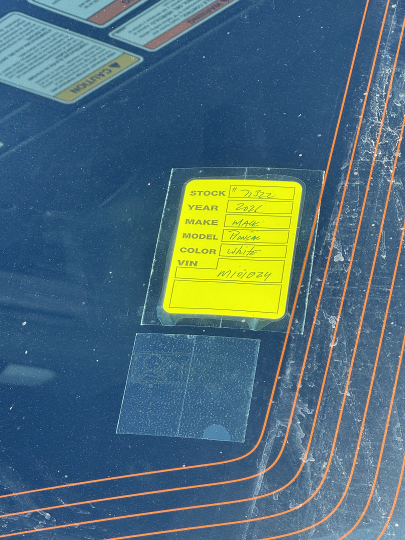 Yellow stock tag on Mack PR64T76H truck showing stock, year, make, model, color, VIN information inside glass window
