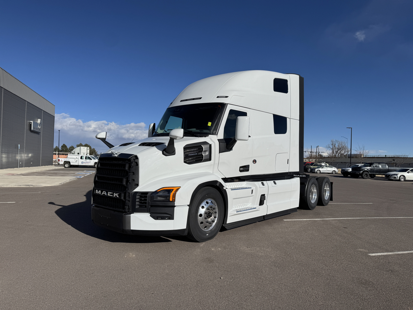 Mack PR64T76H tractor-trailer truck in a parking lot, white cab with sleeper, side view showing front grille and fuel tanks.