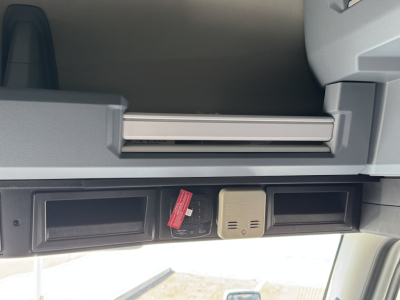 Cab interior overhead shelf with two open cubbies and a beige plug-in device between them in a Mack PR64T76H sleeper cab.