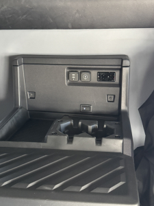 In-cab storage with three rectangular controls on a panel and a small USB/12V port, plus a stepped rubber floor mat.