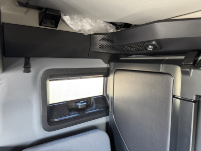 Cab interior of a new Mack PR64T76H sleeper truck showing window with pull-down shade and overhead dashboard area.