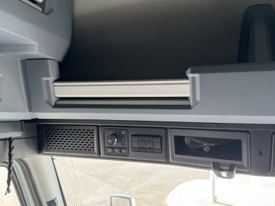 Cab interior dash area of a 2026 Mack PR64T76H showing overhead storage shelf and control panel with vents and empty slot, low lighting