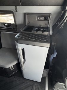 Truck interior with compact fridge and control panel showing USB ports and storage; vertical fridge door and dash area visible.