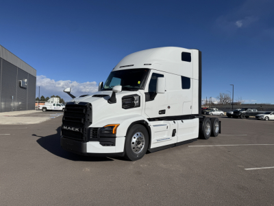Mack PR64T76H tractor-trailer truck in a parking lot, white cab with sleeper, side view showing front grille and fuel tanks.