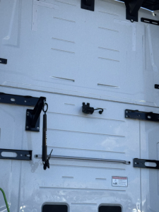 Truck cab interior door area with metal panels and latches showing a small black electrical connector and a long metal rod on a white surface.