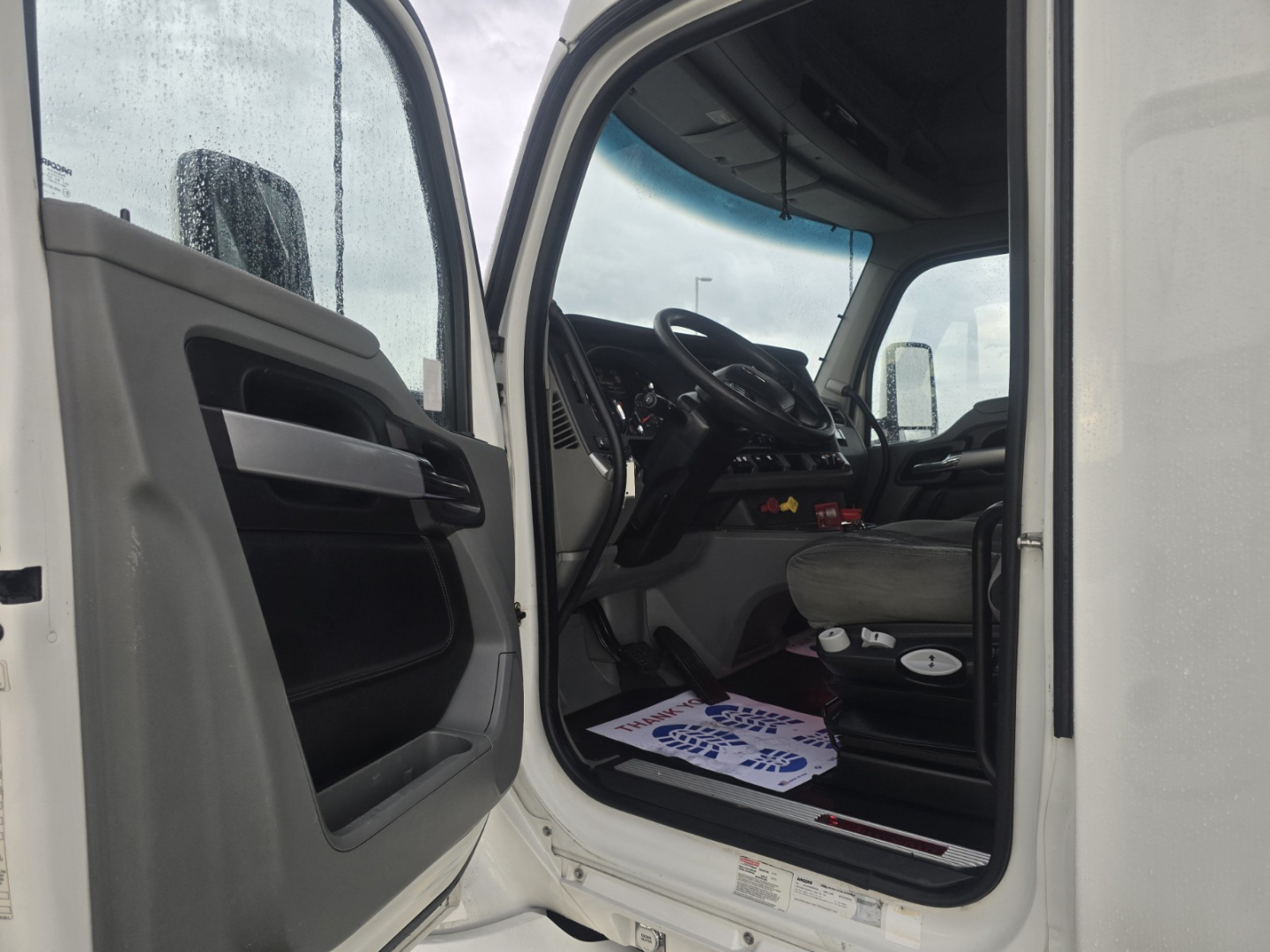 Kenworth T680 truck cab interior view from open door showing steering wheel, dash, and driver's seat; rain on window; plastic floor mat visible.
