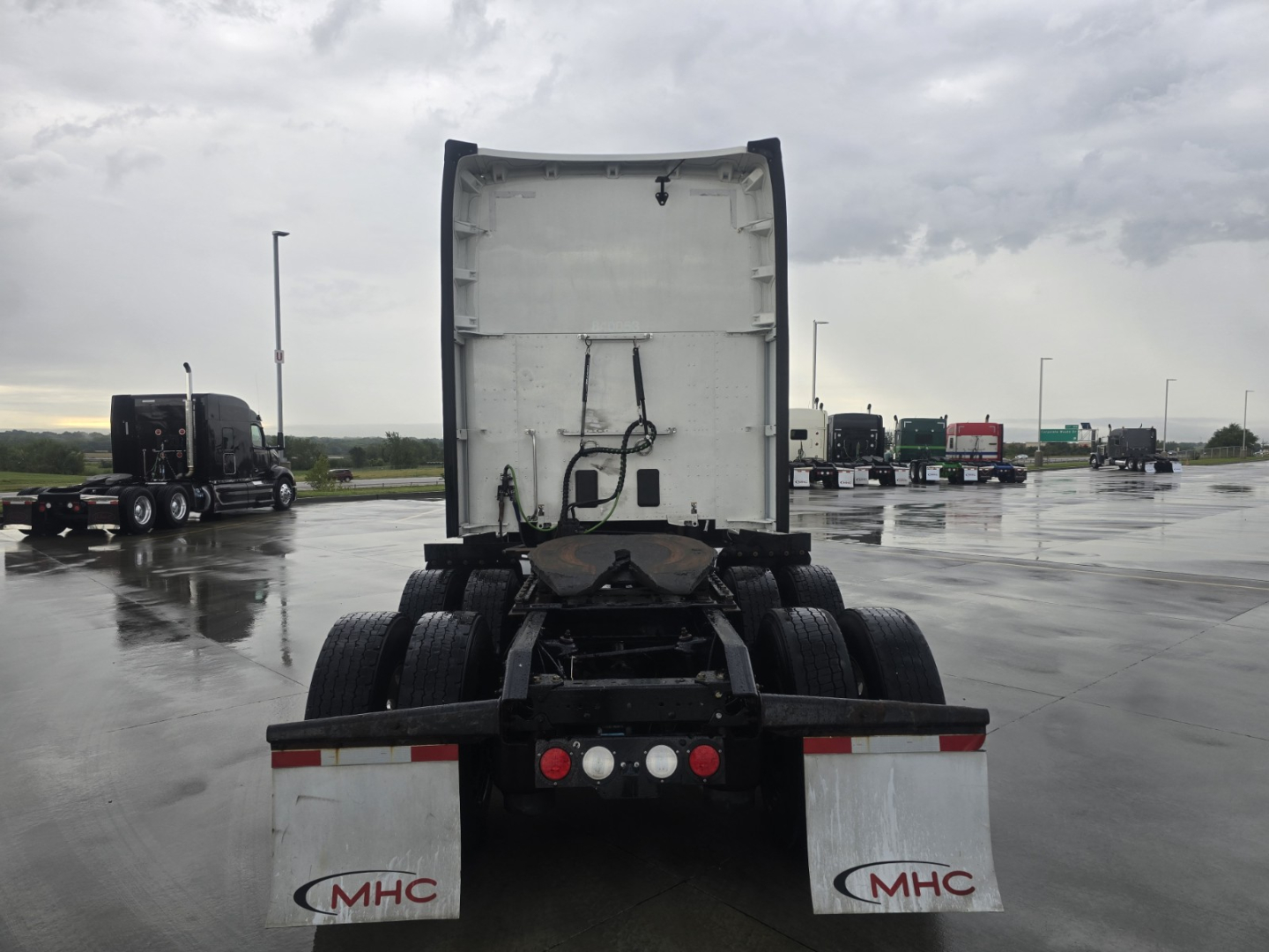 Kenworth T680 rear view showing fifth wheel, rear bumper, mudflaps with MHC logo, and multiple trucks in lot.