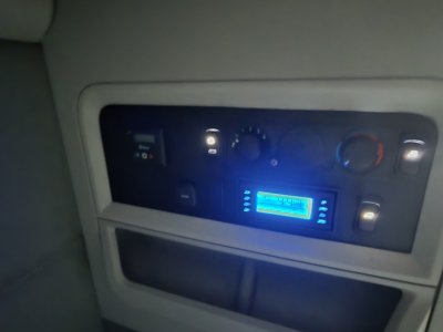 Cabin dashboard panel of a 2021 Kenworth T680 showing control switches, a blue-lit digital radio/display, and gauges.