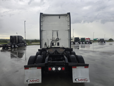 Kenworth T680 rear view showing fifth wheel, rear bumper, mudflaps with MHC logo, and multiple trucks in lot.