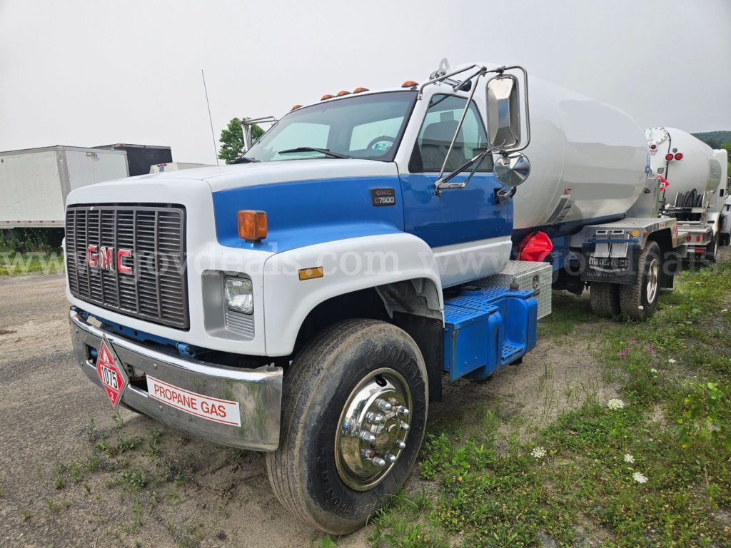 GMC C7500 tanker truck, front three-quarter view, diesel engine bay with propane gas sign, blue and white cab, large front grille, attached cylindrical tanker.